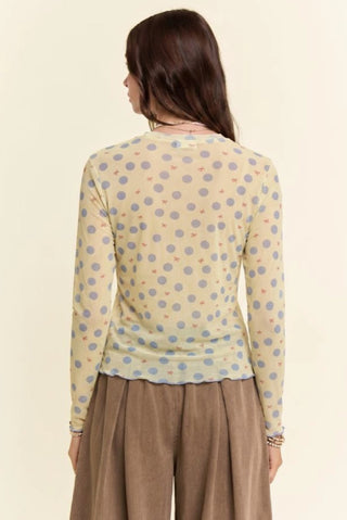 Woman wearing a sheer mesh long-sleeve top with blue polka dots and small red bows on cream background.