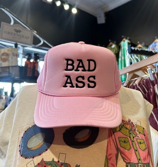 Pink foam trucker hat with black embroidered "BAD ASS" text displayed on mannequin in retail store.