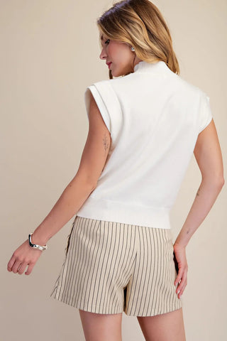 White sleeveless high neck fine knit top with ribbed collar and hem, worn with striped shorts.