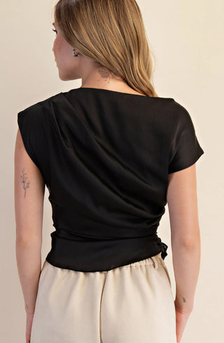 Black boat neck blouse with waist tie detail, short sleeves, worn with cream pants, back view.