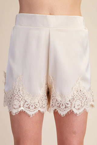 Ivory satin shorts with intricate floral lace trim detail along the hemline, high waist silhouette.