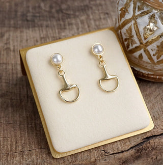 Gold drop earrings featuring round white pearl studs above polished gold equestrian snaffle bit pendants, displayed on cream 