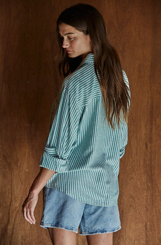 Woman wearing an oversized green and white striped cotton button-front long sleeve collared shirt with denim shorts.