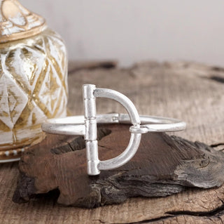 Silver horsebit hook bracelet with snaffle bit design displayed on rustic driftwood with decorative jar.