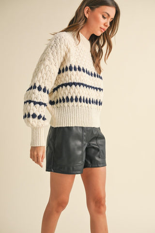 Cream knit sweater with navy blue diamond and stripe pattern, paired with black leather shorts.