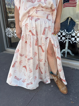 Cream tiered maxi skirt with pink western cowboy boot print, smocked waist, and front slit, styled with tan cowboy boots.