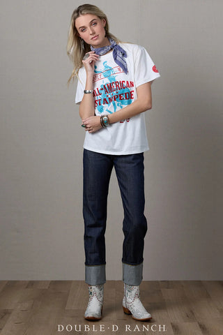 White graphic tee featuring red "All-American Stampede" text and blue rodeo print, styled with dark jeans and cowboy boots.