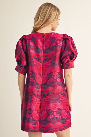 Woman wearing a short magenta and purple jacquard print dress with puff sleeves, shown from behind.
