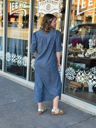Woman wearing a blue chambray midi shirt dress with puff sleeves and ruffle collar, viewed from behind.