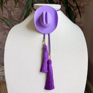 Lavender miniature cowboy hat bolo tie with deep purple tassels and gold metal accents on jewelry display bust.