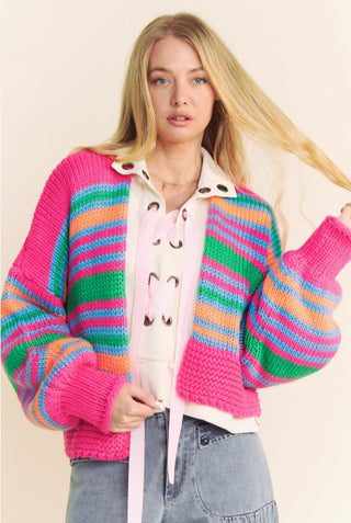 Chunky knit open cardigan with bold multicolor stripes in pink, green, blue, and orange, worn over a cream lace-up top.