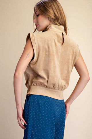 Camel tan suede sleeveless vest with high neck, elastic hem, and structured shoulders, paired with blue polka dot skirt.